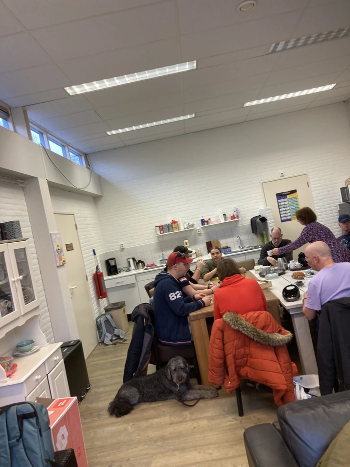 De afbeelding toont een groep mensen die samen aan een tafel zitten in een gemeenschappelijke ruimte. Er zijn zes personen zichtbaar, waarvan er vijf aan tafel zitten en één persoon staat. De ruimte heeft een witte bakstenen muur en een keukenblok met verschillende keukenapparaten en voorwerpen, zoals een koffiezetapparaat en een waterkoker. Op de tafel staan verschillende items, waaronder een thermoskan en wat lijkt op een schaal met eten. Er hangt een rode jas over de rugleuning van een stoel. Op de voorgrond ligt een grote grijze hond ontspannen op de houten vloer. De sfeer lijkt informeel en gezellig.