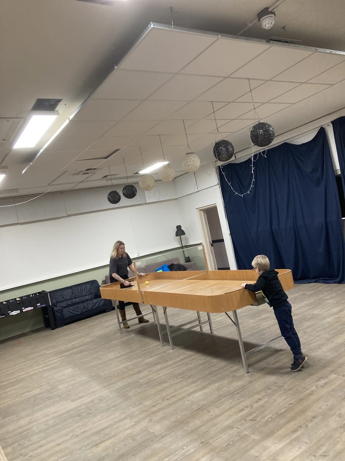 De foto toont een ruime kamer met een houten vloer en een systeemplafond. In het midden van de kamer staat een showdowntafel. Er zijn twee personen te zien: een volwassen vrouw aan de linkerkant van de tafel en een jong kind aan de rechterkant. De vrouw lijkt een showdownbatje vast te houden en is mogelijk bezig met het spelen van een spel. Het kind kijkt naar de tafel en lijkt ook betrokken te zijn bij het spel. Aan het plafond hangen decoratieve bollen in zwart en wit, en er is een donkerblauw gordijn aan de rechterkant van de kamer. Links in de kamer staat een zwarte bank tegen de muur