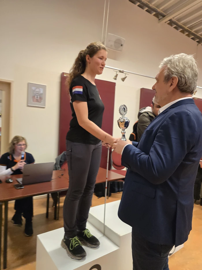 Op de foto staat een vrouw op een podium met het nummer 2 erop. Ze draagt een zwart T-shirt met het Cannonballs-logo en een Nederlandse vlag op de mouw en een donkergrijze broek. Ze heeft lang, golvend haar en draagt zwarte sportschoenen met felgroene veters. Ze ontvangt een trofee van een man in een donkerblauw colbert. De man heeft grijs haar en een baard. Op de achtergrond zit een vrouw achter een tafel met een laptop. 