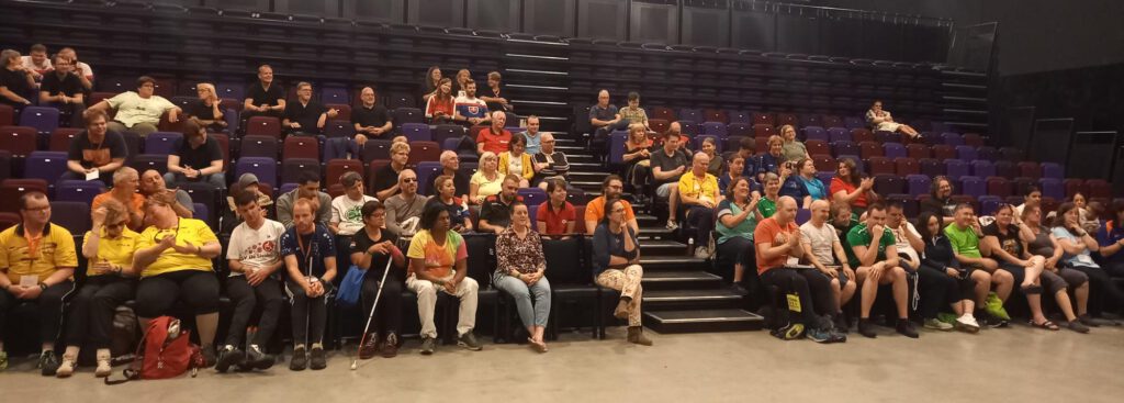 Publiek in een zaal waarbij mensen op kleurige stoelen in tribunes zitten. De sfeer is ontspannen en iedereen kijkt richting het podium, in afwachting op wat er gaat komen. Aan de voorzijde zitten mensen op de eerste rij, sommigen in sportkleding, anderen in informele kleding. Er is een mix van leeftijden en achtergronden zichtbaar. Enkele mensen praten, anderen lachen of kijken geconcentreerd naar voren. De zaal is goed gevuld, vooral in het midden en vooraan.